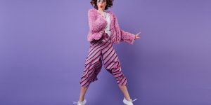 Full-length shot of glad curly woman in striped pants jumping on purple background. Indoor portrait of wonderful girl in sunglasses fooling around in studio.